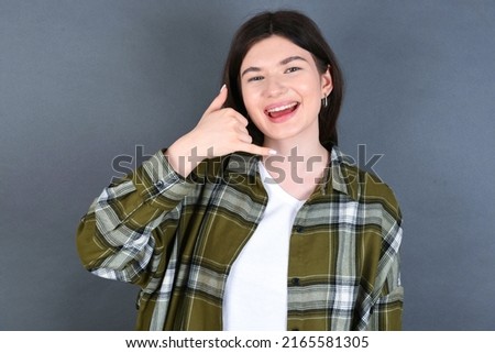 young beautiful Caucasian woman wearing overshirt over studio grey wall, makes phone gesture, says call me back again, has glad expression.