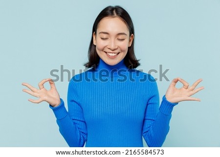 Young woman of Asian ethnicity 20s years old wears blue shirt hold spreading hands in yoga om aum gesture relax meditate try to calm down isolated on plain pastel light blue background studio portrait Royalty-Free Stock Photo #2165584573