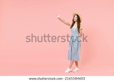 Full length young cheerful happy woman she in summer clothes striped dress straw hat doing selfie shot on mobile cell phone show victory gesture v-sign isolated on pastel pink color background studio