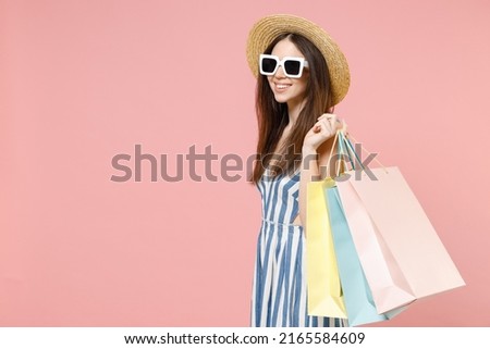 Side view young happy excited caucasian woman wearing summer clothes striped dress straw hat glasses hold package bags with purchases after shopping isolated on pastel pink background studio portrait