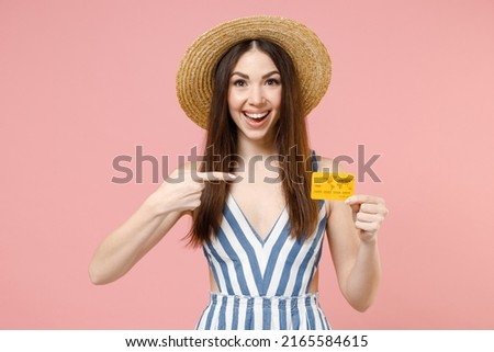 Young fun caucasian rich smiling happy woman 20s in summer clothes striped dress straw hat put index finger on credit bank card looking camera isolated on pastel pink color background studio portrait.
