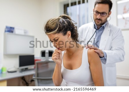 Young female patient in the clinic suffered from pneumonia, she is coughing the doctor listens to the wheezing in the lungs with a stethoscope. Royalty-Free Stock Photo #2165596469