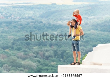 Dad and son are standing on observation deck holding hand above eyes and peering into distance. Boy sits on the mans shoulders