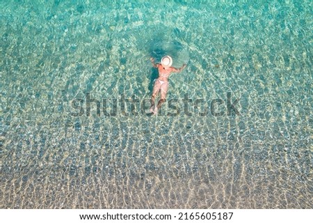 Top, aerial view. Young beautiful woman in a hat and white bikini swimming in sea water on the sand beach. Drone, copter photo. Summer vacation. View from above. 
