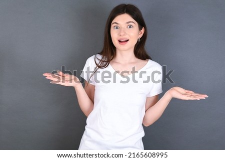 So what? Portrait of arrogant young beautiful Caucasian woman wearing white T-shirt over studio grey wall, shrugging hands sideways smiling gasping indifferent, telling something obvious. Royalty-Free Stock Photo #2165608995