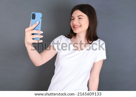 Isolated shot of pleased cheerful young beautiful Caucasian woman wearing white T-shirt over studio grey wall,, makes selfie with mobile phone. People, technology and leisure concept