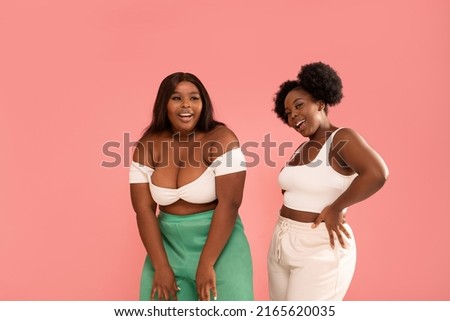 Happy dark skinned, young sisters enjoying good moment, dancing and having fun together, posing over pink pastel studio background. Friendship and real people emotions concept. Body positive.