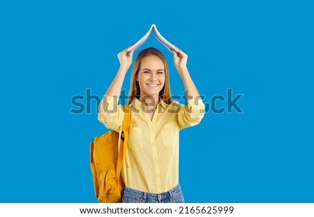 Portrait of smiling Caucasian teenage girl with backpack hold book over head excited about studying. Happy millennial female student with textbook isolated on blue studio background. Education.