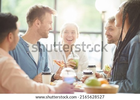 Group of joyful young multi-ethnic friends in casual clothing sitting at one table and eating pizza while chatting during lunch