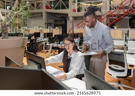 two multiracial coworkers sitting at a table in a coworking space working together and looking at a computer screen and papers.