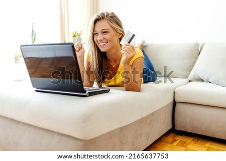 Happy Caucasian young woman lying on sofa with laptop computer and credit card at home. Smiling young woman with laptop computer and credit card. Online shopping, banking and technology concept.