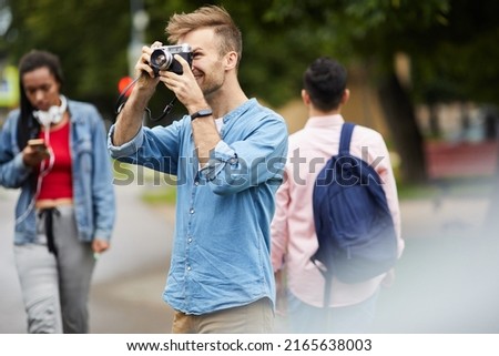 Smiling handsome young man in blue shirt standing in city park and photographing nature on old camera