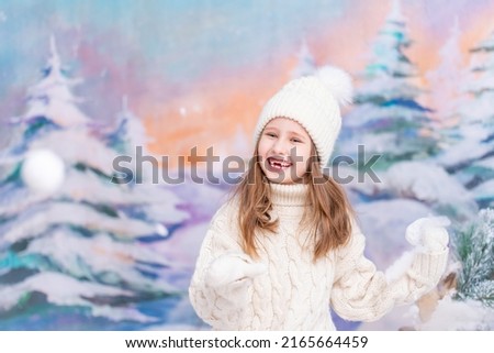 happy blue-eyed girl-child in a hat and a knitted sweater, playing snowballs throwing them up, stands against the background of a winter forest. Winter holidays, warm clothes