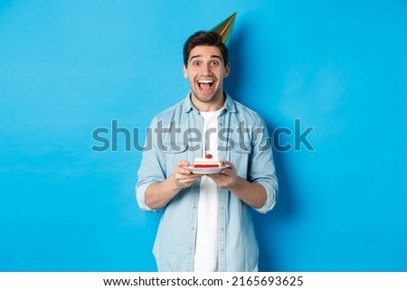 Cheerful young man celebrating birthday in party hat, holding b-day cake, standing against blue background
