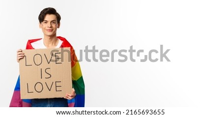 Smiling gay man activist holding sign love is love for lgbt pride parade, wearing Rainbow flag, standing over white background