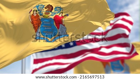 The New Jersey state flag waving along with the national flag of the United States of America. In the background there is a clear sky. New Jersey s a state in the Northeastern regions of the US Royalty-Free Stock Photo #2165697513