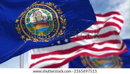 The New Hampshire state flag waving along with the national flag of the United States of America. In the background there is a clear sky. New Hampshire is a state in the New England region of the US Royalty-Free Stock Photo #2165697515
