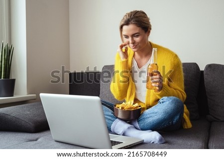 Young woman is sitting on the sofa drinking beer and eating nachos while watching movie on her laptop at home Royalty-Free Stock Photo #2165708449