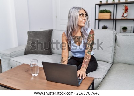 Middle age grey-haired woman using laptop at home looking to side, relax profile pose with natural face with confident smile. 