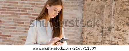young redhead woman using smartphone on european street near brick wall, banner