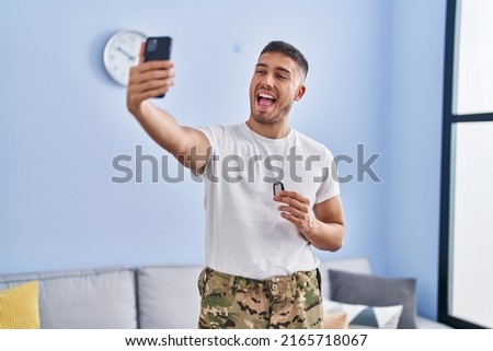 Young hispanic man wearing camouflage army uniform taking selfie at home smiling and laughing hard out loud because funny crazy joke. 