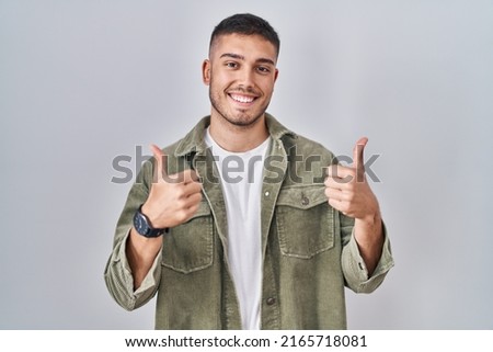 Young hispanic man standing over isolated background success sign doing positive gesture with hand, thumbs up smiling and happy. cheerful expression and winner gesture.  Royalty-Free Stock Photo #2165718081