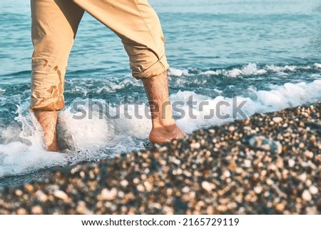 Man wearing shorts, walking barefoot along the seashore. Male legs walks on pebble beach along the shore near the water with waves, low section. Wellness, freedom and travel in summertime concept. Royalty-Free Stock Photo #2165729119
