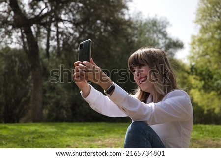Portrait of young, beautiful, blonde woman in white shirt and jeans, taking a selfie with her cell phone in the middle of a park. Mobile concept, selfie, photo, portrait, technology.