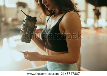 Cropped picture of a strong sportswoman sits in a gym, drinks protein drink and using a phone while drinking protein drink and taking a break.