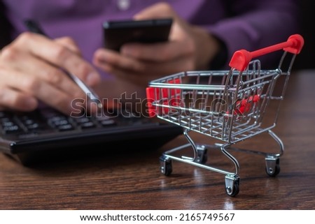 Online shopping concept. Small supermarket grocery push cart for shopping, A man counts the cost of purchases on a calculator.