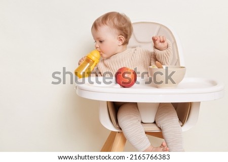 Image of thirsty toddler baby girl dresses in beige jumper sitting in high chair and drinking water from yellow bottle, looking aside, studying area around her, posing isolated over white background Royalty-Free Stock Photo #2165766383