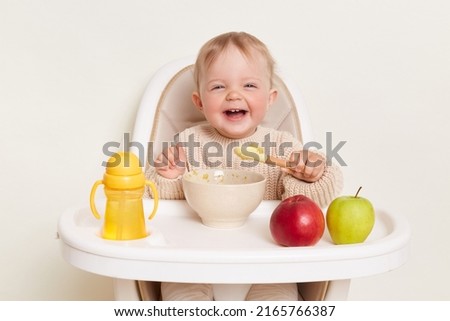 Image of deligted laughing infant baby girl dresses in beige jumper sitting in high chair and eating, isolated over white background, holding spoon in hands, enjoying tasty fruit puree. Royalty-Free Stock Photo #2165766387