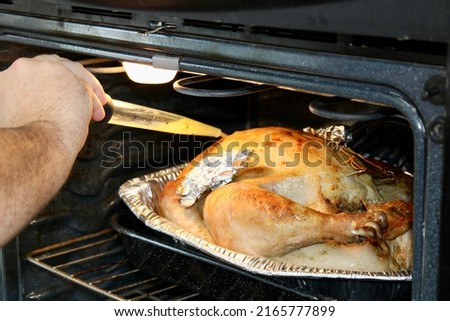 A man basting a whole turkey that is baking in the oven. The giant turkey has it's wings wrapped in tin foil.