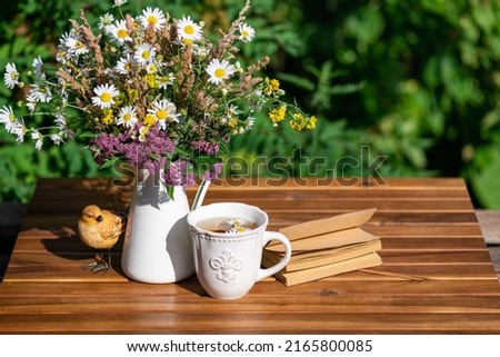 Garden composition bouquet of meadow flowers, herbal tea cup, books on table in summer. Rest in garden, reading books, breakfast, rest in nature concept. Summertime in garden on backyard