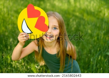 Sweet blue-eyed little girl is holding a cardboard love emoticon with hearts in hands. A happy child on a natural background