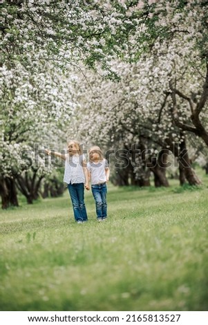 Cute children stand in a blooming garden and wipe off white petals. Beautiful sisters are walking in the park among a blooming garden.