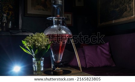 Magic hookah smoke in vintage interior with small candle and bouquet of lilies of the valley. Design glass hookah with metal frame. Illuminated by red lights. Space for text.