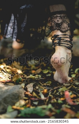 Beautiful feet of young gothic and witch woman with black nails and accessories walking over autumn brown and yellow leaves in the forest ground
