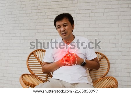 Middle aged, senior Asian man with sudden onset of heart disease showing pain heart muscle leaks tired and suffocating sits on a wooden chair in agony : Myocardial infarction (Heart Attack) Royalty-Free Stock Photo #2165858999