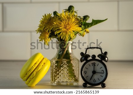 Macaroons and a vase with yellow dandelions and an alarm clock. Good morning. French pasta on the table and a bouquet of yellow dandelions in a vase. Selective focus.