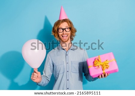Photo of cute dreamy young guy dressed jeans shirt eyewear headwear holding balloon present empty space isolated blue color background