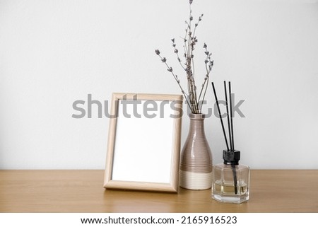 Blank frame, vase with flowers and reed diffuser on table near light wall