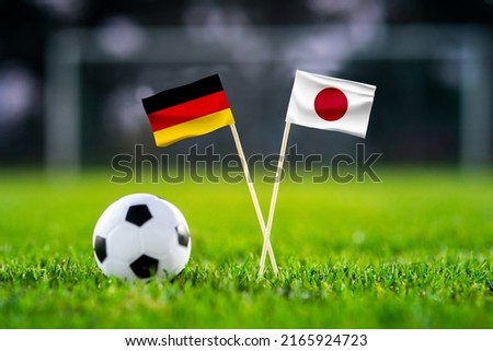 October 2022: Germany vs. Japan, Khalifa Stadium, Football match wallpaper, Handmade national flags and soccer ball on green grass. Football stadium in background. Black edit space.
