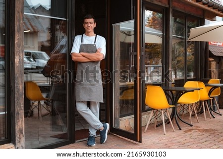 Portrait barista or waiter cafe or coffee shop owner against entrance, gesture inviting you to visit, smiling guy in apron standing outdoors being proud of his small local business Royalty-Free Stock Photo #2165930103