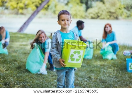 Happy boy holding a recycling bin while cleaning the surroundings with his friends and looking at the camera. An adorable preschool age little boy stands outdoors in a public park.