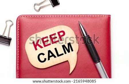 On a light background there are black paper clips, a pen, a burgundy notepad a wooden board with the text KEEP CALM