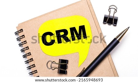 On a light background, a brown notebook, a black pen and paper clips, and a yellow card with the text CRM Customer Relationship Management