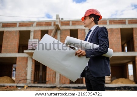 Young man engineer architect in red helmet is holding a paper plan of a building at the construction site.
