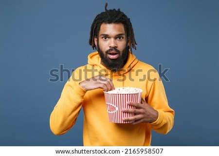 Amazed scared young man of African American ethnicity 20s he wears casual yellow hoodie watch movie film in cinema hold bucket of popcorn isolated on plain dark blue royal background studio portrait