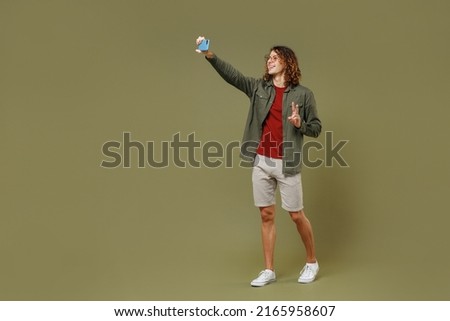 Full size body length side view young brunet curly man 20s wears khaki shirt do selfie shot on mobile cell phone post photo on social network isolated on plain olive green background studio portrait
